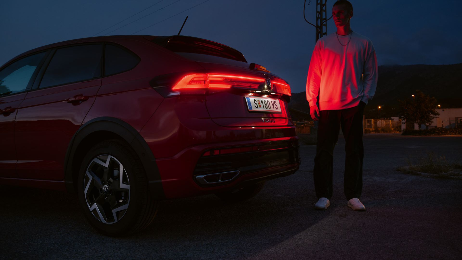 Ein Mann steht am Heck eines VW Taigo R-Line im Dunkeln und wird von den Rückleuchten rot angeleuchtet.