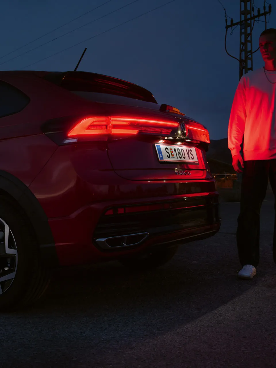 Ein Mann steht am Heck eines VW Taigo R-Line im Dunkeln und wird von den Rückleuchten rot angeleuchtet.