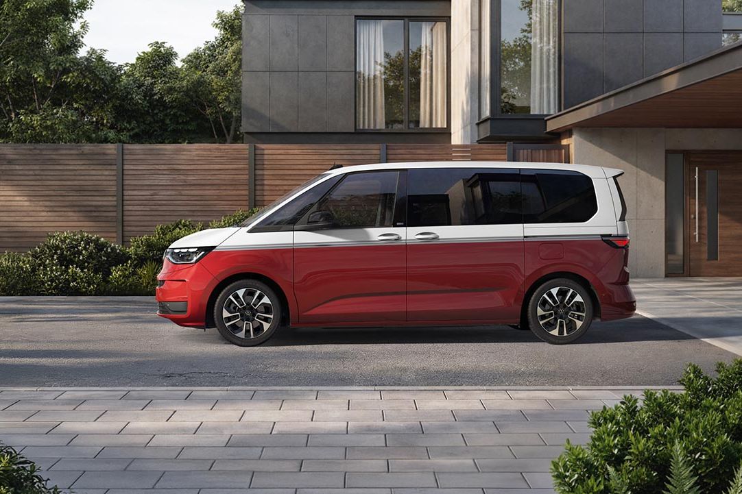 Ein rot weißer Volkswagen Multivan steht seitlich auf einer gepflasterten Einfahrt vor einem schicken, minimalistischen Wohnhaus.
