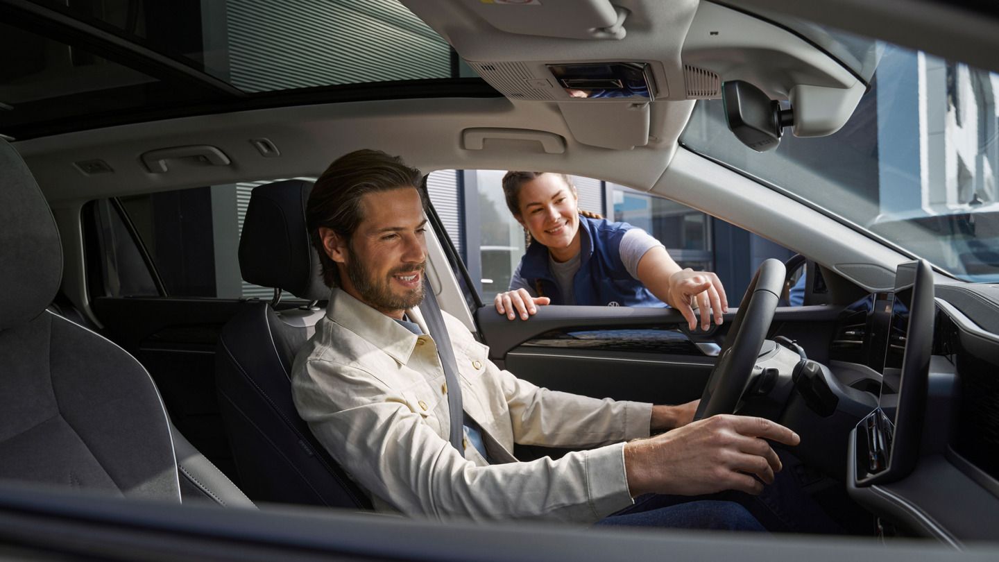 Eine VW AService Mitarbeiterin zeigt einem Kunden, welcher im Auto sitzt die neuen Funktionen im Volkswagen.