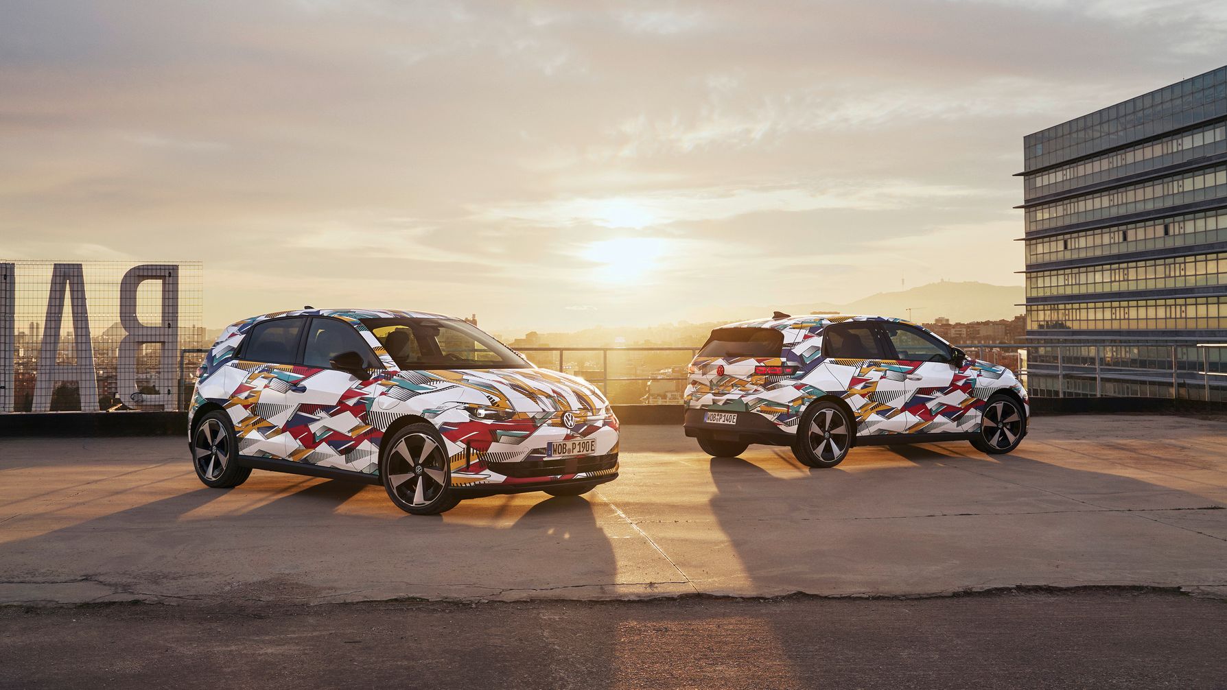 Zwei VW ID. Polos mit bunter Folierung stehen nebeneinander auf einem Parkplatz im Freien bei Sonnenuntergang. Im Hintergrund sind moderne Bürogebäude zu sehen. Die Sonne geht am Horizont unter und beleuchtet die Szene.