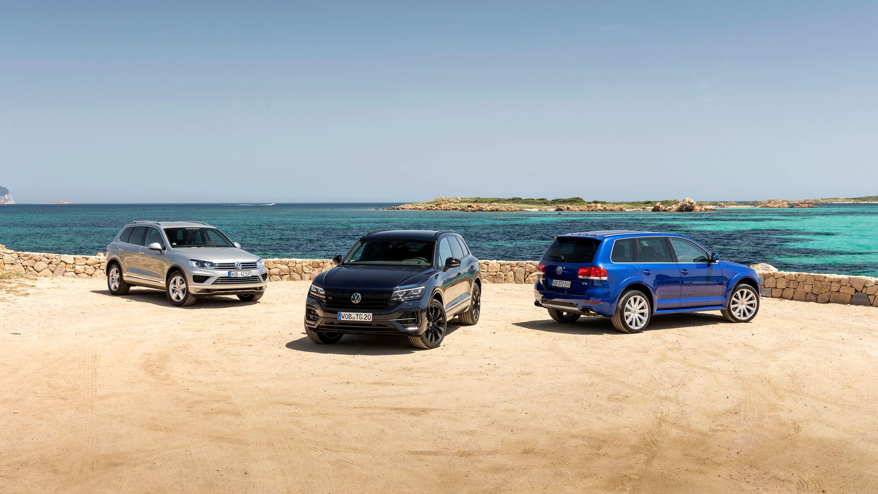 Drei VW Touareg stehen zusammen an einem Strandparkplatz am Meer.