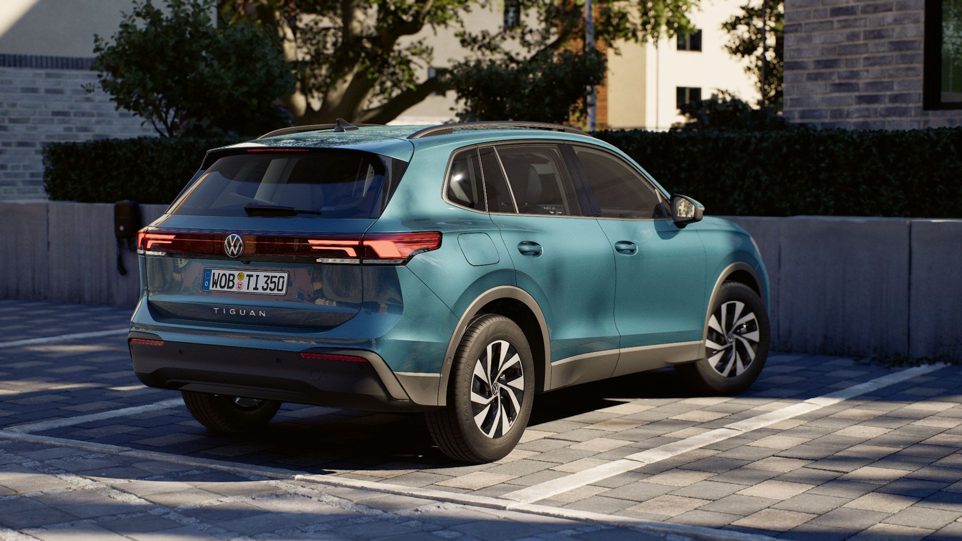 Ein blauer Tiguan steht rückwärts in einer markierten Parklücke auf einem gepflasterten Parkplatz. Das Auto ist modern gestaltet und wird leicht von hinten schräg gezeigt, sodass Heckleuchten, Seitentür und ein Teil der Front sichtbar sind. Im Hintergrund befinden sich eine niedrige Mauer, Büsche und Gebäudefassaden in warmem Sonnenlicht.