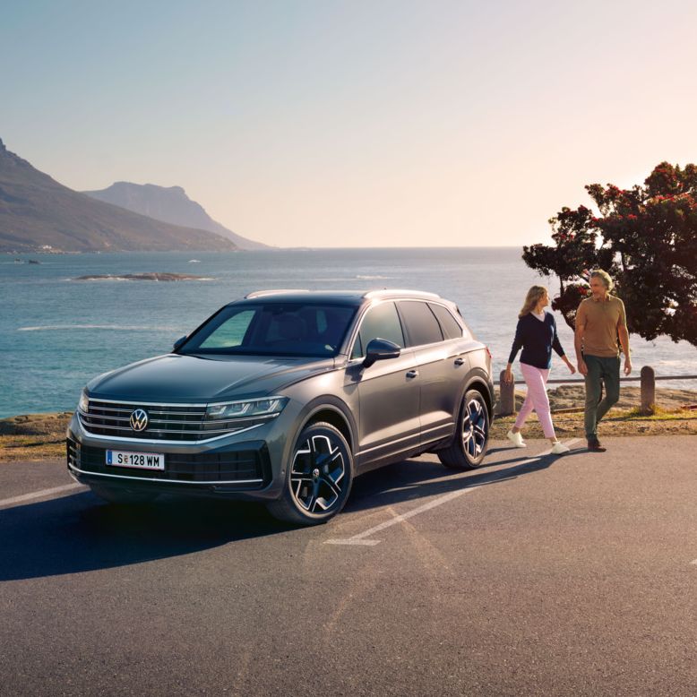 Der neue Volkswagen Touareg auf einem Parkplatz an der Küste, an ihm gehen eine Frau und ein Mann vorbei