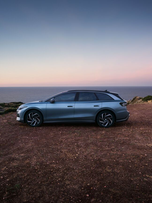 Ein blauer VW ID.7 Tourer steht oben auf einer Klippe im Sonnenuntergang, dahinter das Meer.