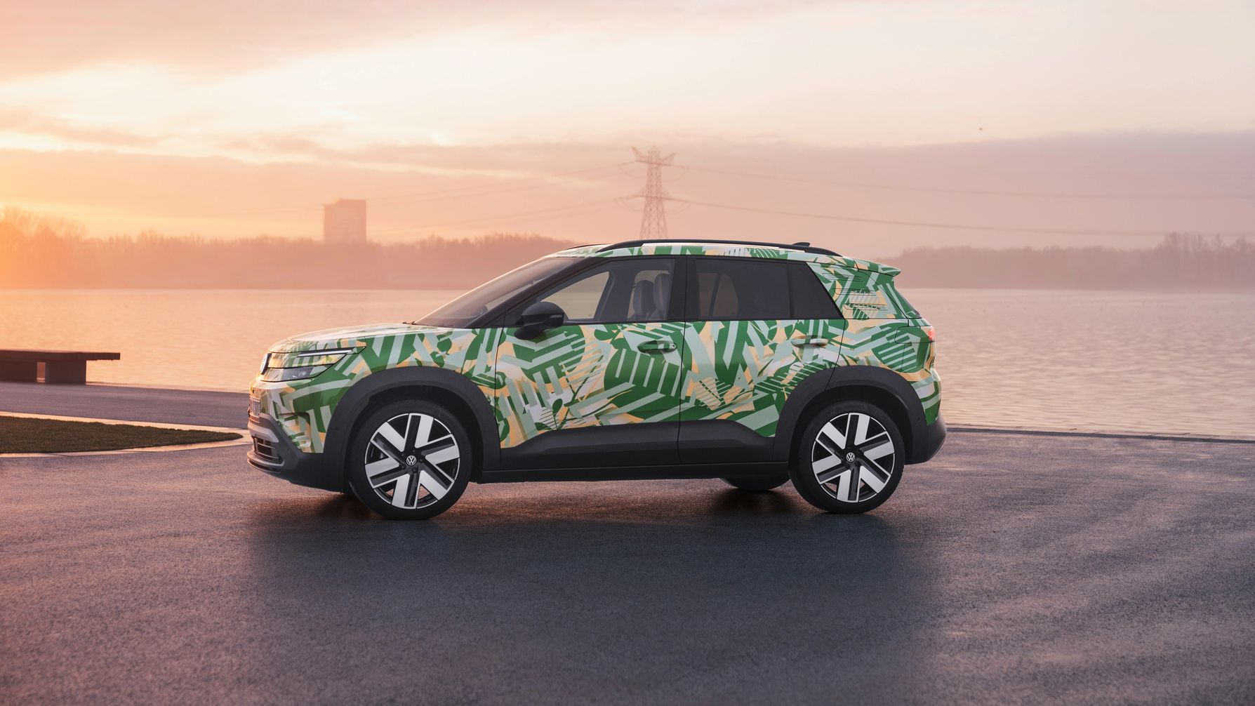 Ein SUV mit einer grünen und weißen Tarnlackierung steht auf einem asphaltierten Platz vor einem Gewässer bei Sonnenuntergang. Im Hintergrund sind Strommasten und Gebäude zu sehen. Der Himmel ist leicht bewölkt und in warmen Farben gehalten.