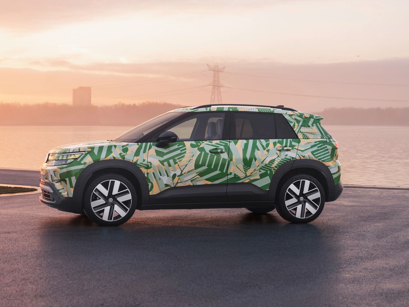 Ein SUV mit einer grünen und weißen Tarnlackierung steht auf einem asphaltierten Platz vor einem Gewässer bei Sonnenuntergang. Im Hintergrund sind Strommasten und Gebäude zu sehen. Der Himmel ist leicht bewölkt und in warmen Farben gehalten.