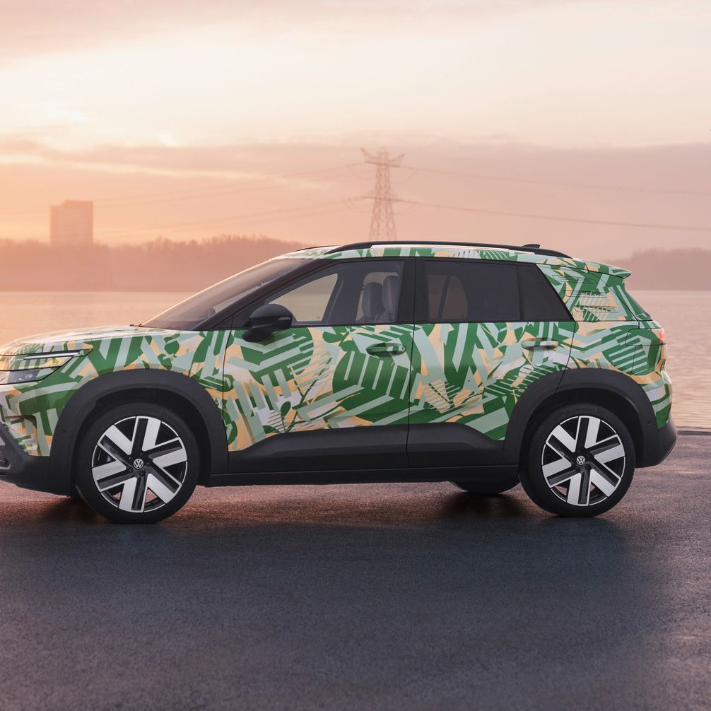 Ein SUV mit einer grünen und weißen Tarnlackierung steht auf einem asphaltierten Platz vor einem Gewässer bei Sonnenuntergang. Im Hintergrund sind Strommasten und Gebäude zu sehen. Der Himmel ist leicht bewölkt und in warmen Farben gehalten.