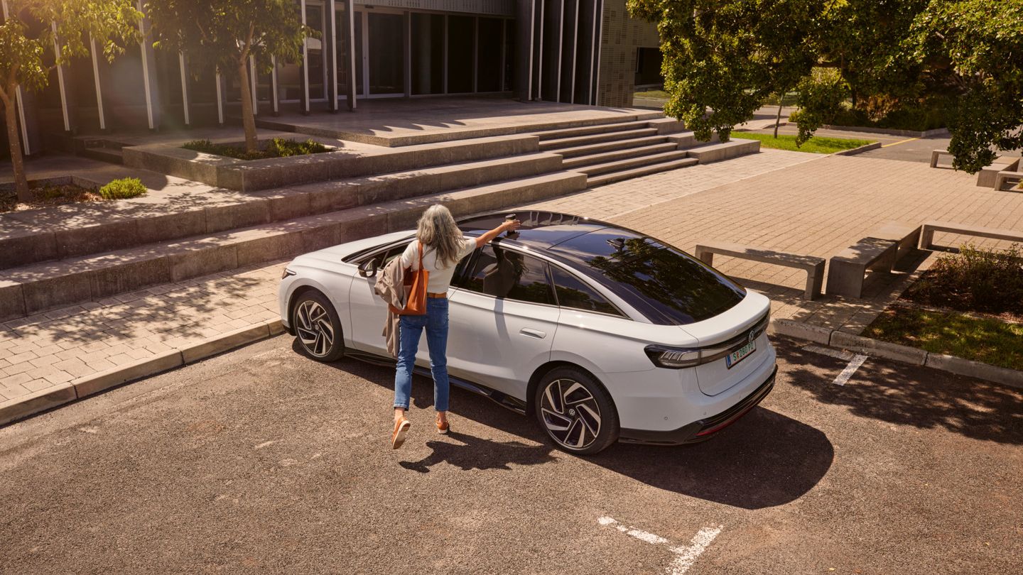 Eine Frau nimmt einen Becher vom Dach eines VW ID.7, der vor einem Bürogebäude parkt.