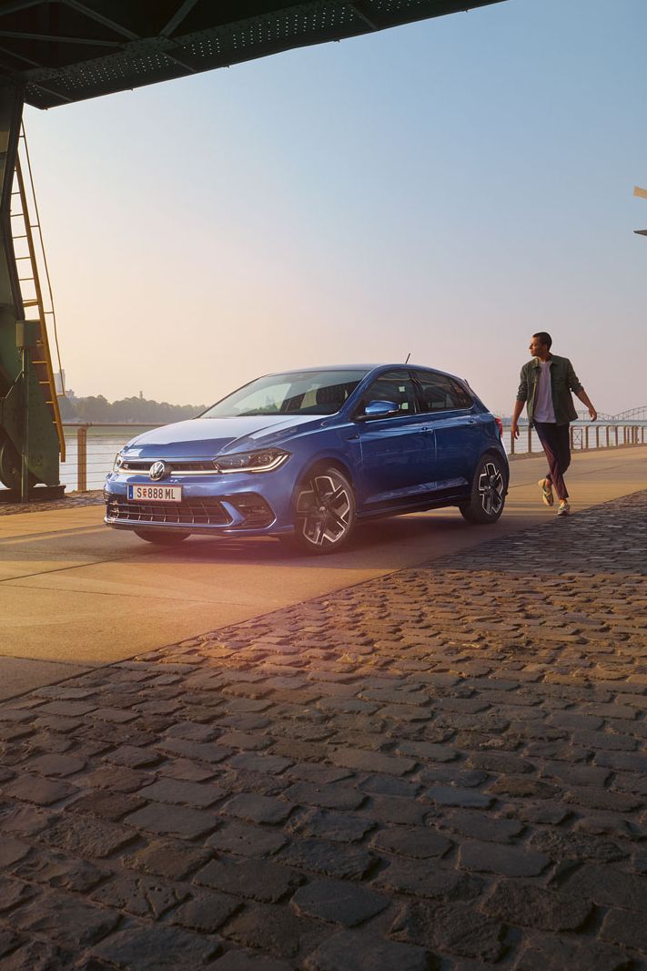  Ein blauer VW Polo R-Line steht an der Promenade neben einem Fluss, ein Mann läuft um das Auto herum.
