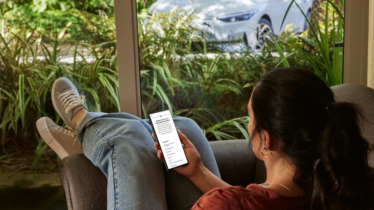 Eine Frau sitzt entspannt im Sofa und blickt auf ihr Smartphone, im Hintergrund ein VW ID.7
