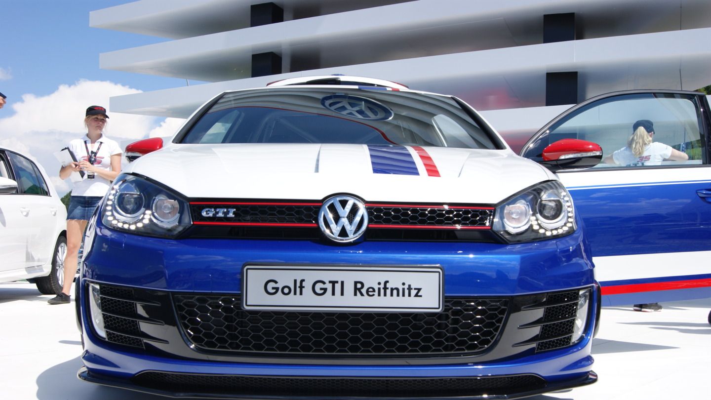 Ein blauer VW Golf GTI mit roten und weißen Akzenten steht vor einem modernen Gebäude, im Hintergrund sind weitere Autos zu sehen.