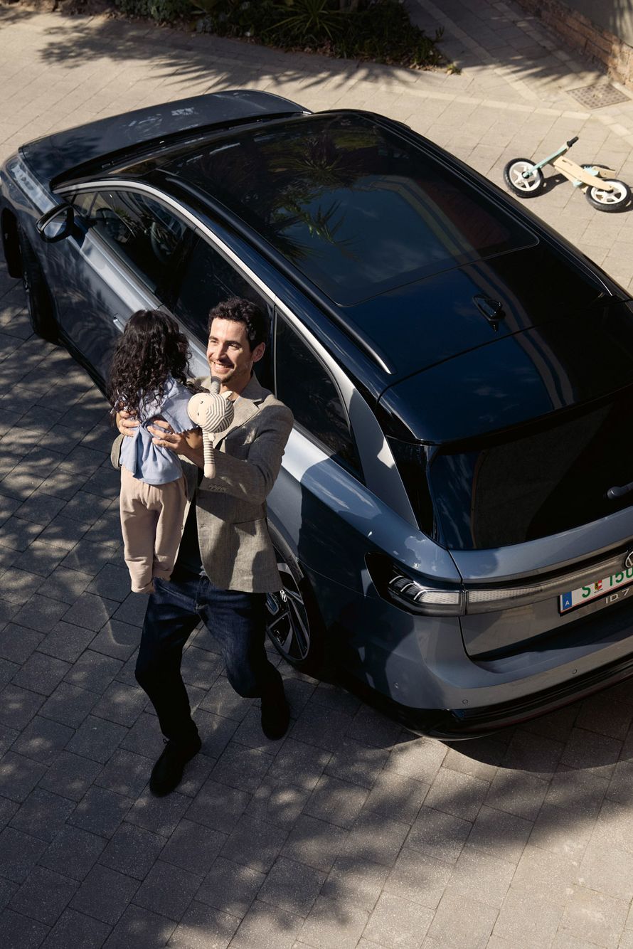 Mann hebt sein Kind hoch, was eine Puppe in der Hand halt und dahinter steht geparkt in der Einfahrt ein ID.7 Tourer und ein umgekipptes Kinderfahrrad.