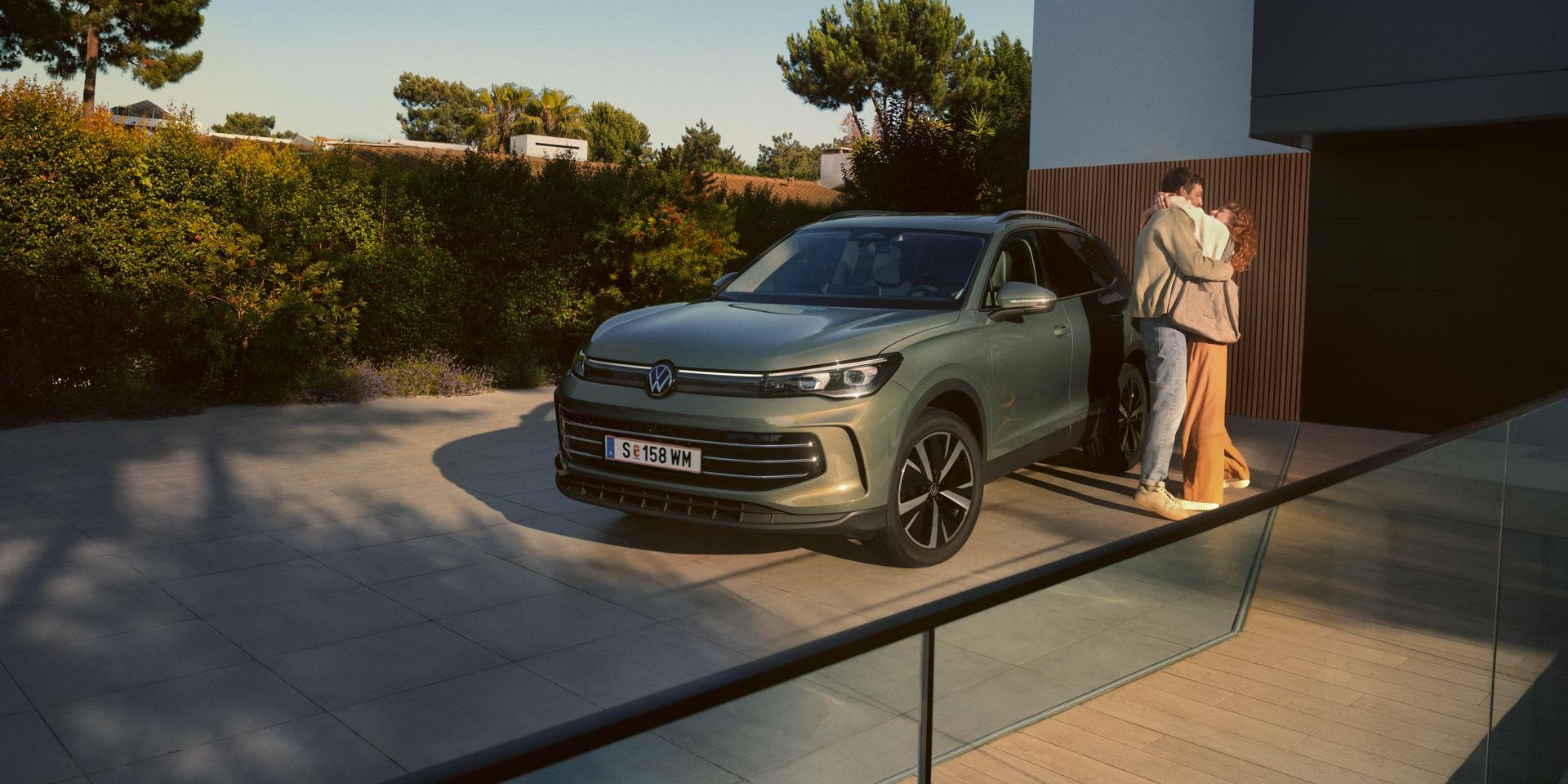 Grüner VW Tiguan parkt in Frontansicht auf der Einfahrt eines modernen Wohnhauses. Ein junges Paar steht daneben und umarmt sich.
