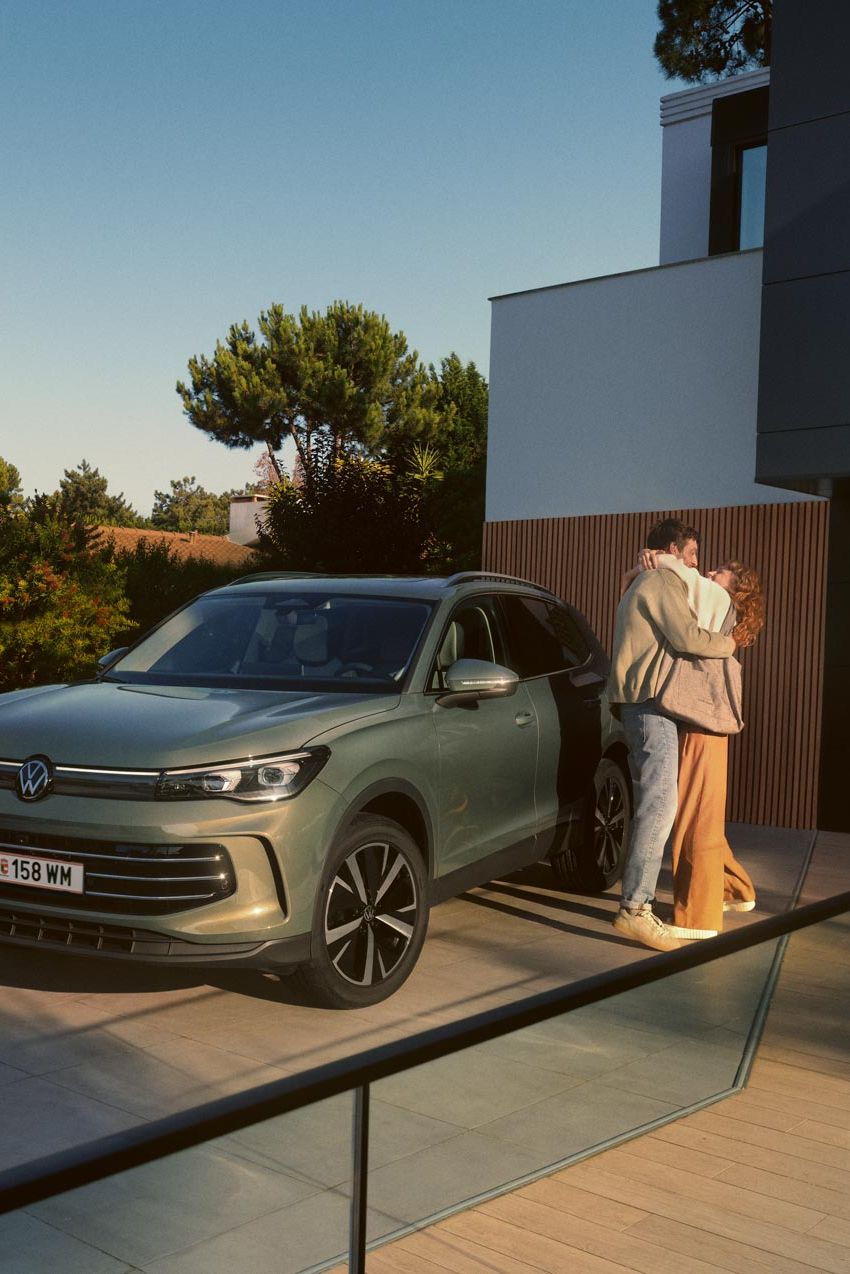 Grüner VW Tiguan parkt in Frontansicht auf der Einfahrt eines modernen Wohnhauses. Ein junges Paar steht daneben und umarmt sich.
