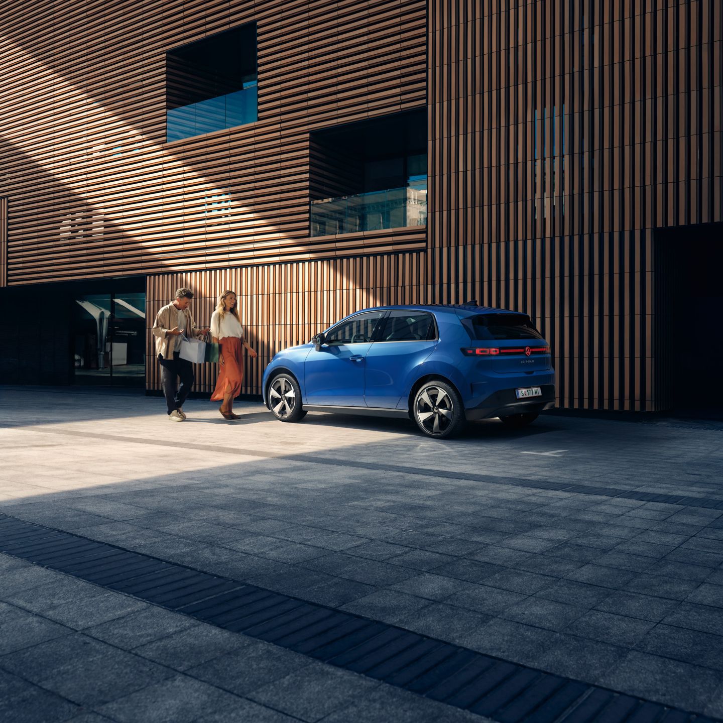 Ein blauer VW ID. Polo Style steht vor einem Gebäude mit Holzfassade. Ein Mann und eine Frau mit Einkaufstaschen gehen darauf zu