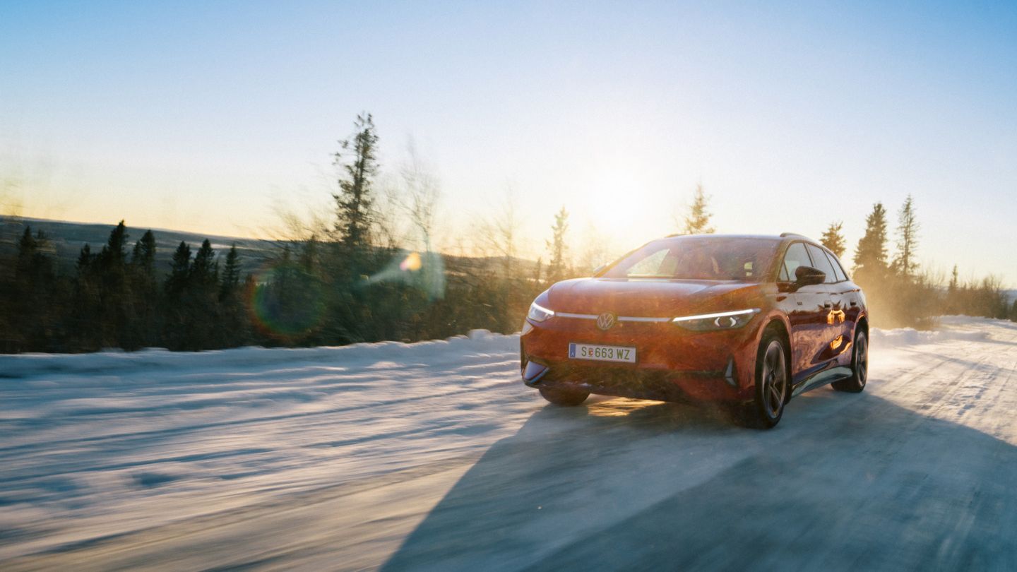Ein roter VW ID.7 Tourer GTX fährt bei Sonnenuntergang auf einer Landstraße. Die untergehende Sonne wirft ein warmes, goldenes Licht auf die Umgebung und das Fahrzeug.