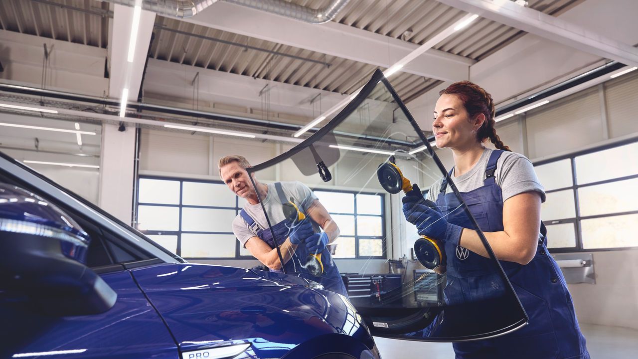 Zwei Volkswagen-Monteur:innen in blauen Overalls setzen mit Saugnapfwerkzeugen eine große Autoscheibe in einen blauen Volkswagen ein.