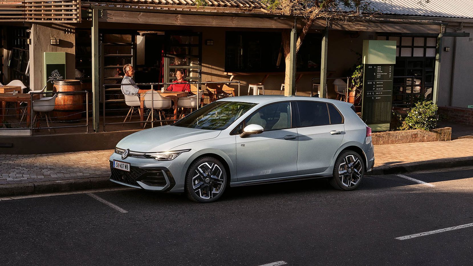 Ein Paar sitzt auf einer Restaurantterrasse, davor parkt ein weißer VW Golf.