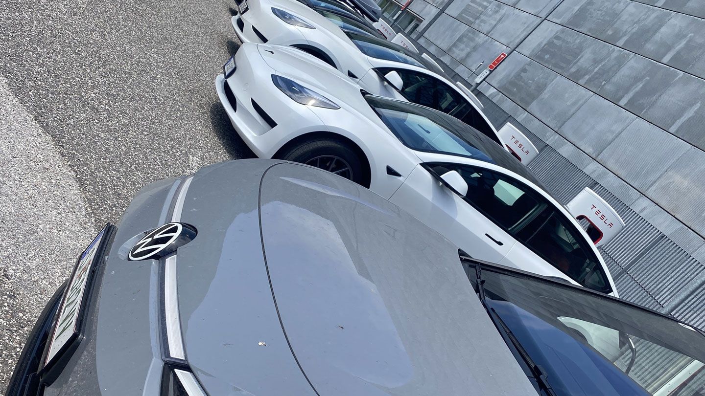Mehrere Elektroautos, größtenteils Tesla-Modelle, sind nebeneinander in einer Reihe auf einem Parkplatz abgestellt. Die Autos sind weiß und grau. Im Hintergrund befindet sich ein Gebäude mit Tesla-Schildern.