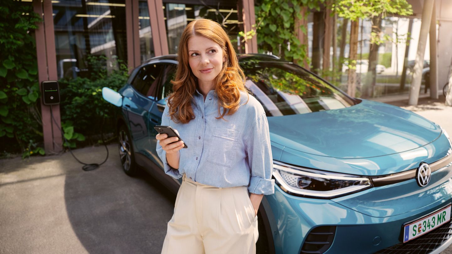 Frau mit Smartphone in der Hand steht vor der Motorhaube eines blauen VW ID.4, der an einer Wallbox angeschlossen ist.