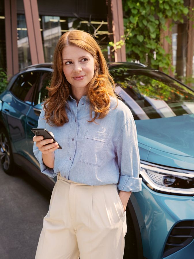Frau mit Smartphone in der Hand steht vor der Motorhaube eines blauen VW ID.4, der an einer Wallbox angeschlossen ist.