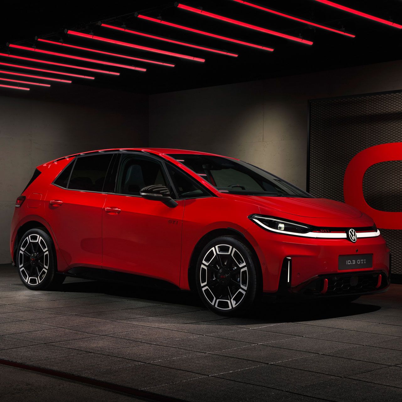 Roter, sportlich wirkender Elektro-Kompaktwagen in einem dunklen Showroom mit roten Neon-Deckenleuchten. Das Auto steht im Profil leicht nach rechts gedreht, mit leuchtenden LED-Scheinwerfern und großen schwarzen Felgen mit Zierakzenten. Rechts im Hintergrund ist eine Gitterwand mit großem roten Schriftzug zu sehen, die die dynamische, sportliche Szene unterstreicht.