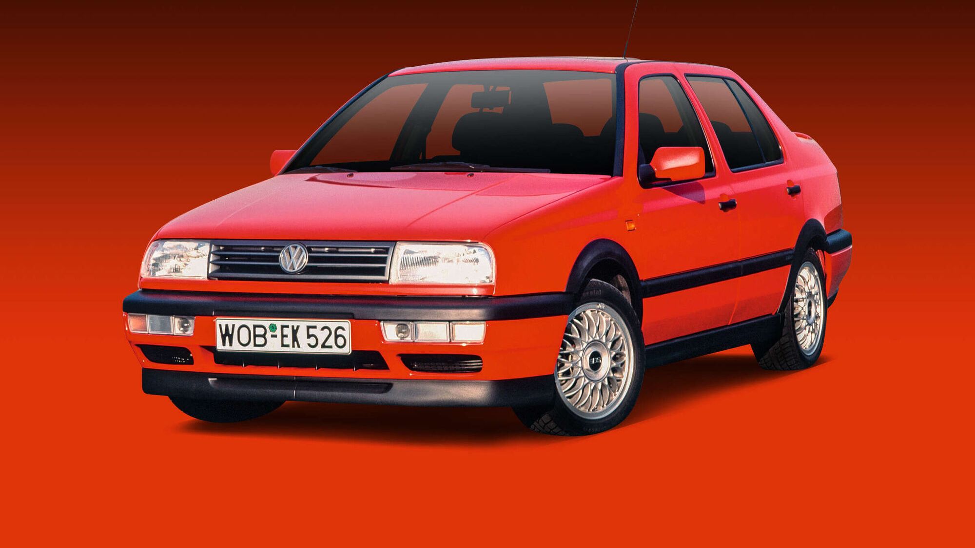 Ein roter VW Jetta der 1990er-Jahre ist vor einem Verlaufshintergrund in Rottönen freigestellt abgebildet. Das Auto steht leicht schräg nach rechts gedreht, sodass Front und Fahrerseite gut sichtbar sind. Details wie der VW-Logo-Grill, die eckigen Scheinwerfer und die mehrspeichigen Alufelgen sind klar erkennbar.
