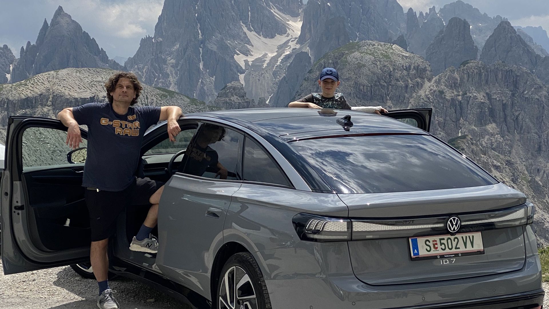 Zwei Personen lehnen an einem grauen Volkswagen ID.7, der vor einer beeindruckenden Berglandschaft mit schroffen Gipfeln steht. Der Himmel ist leicht bewölkt. Die Stimmung ist entspannt und abenteuerlich.
