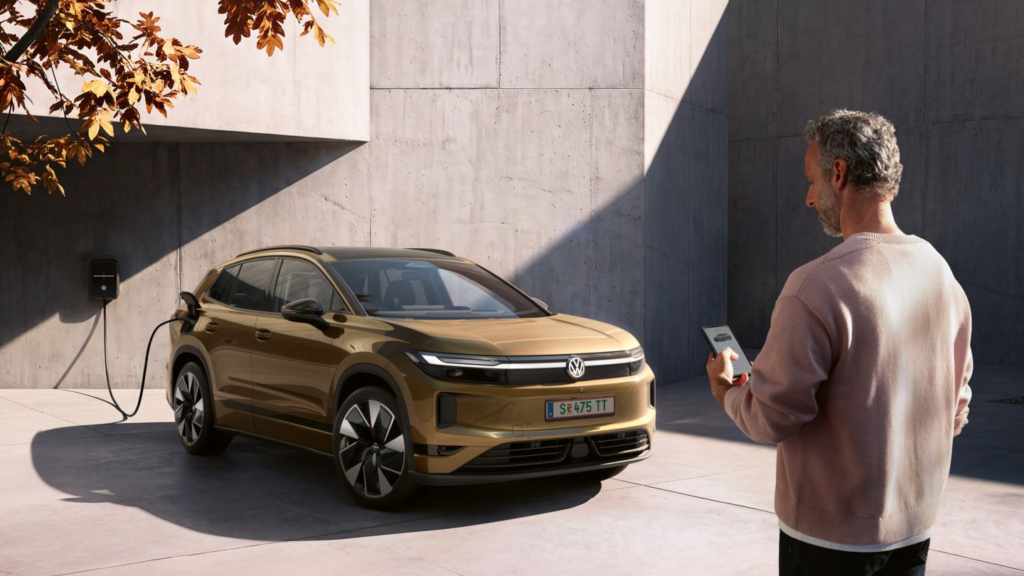 Ein Mann schaut auf das Handy in seiner Hand. Im Hintergrund lädt ein oliv-bronzefarbener VW ID.4 Reskin an einer Wallbox.