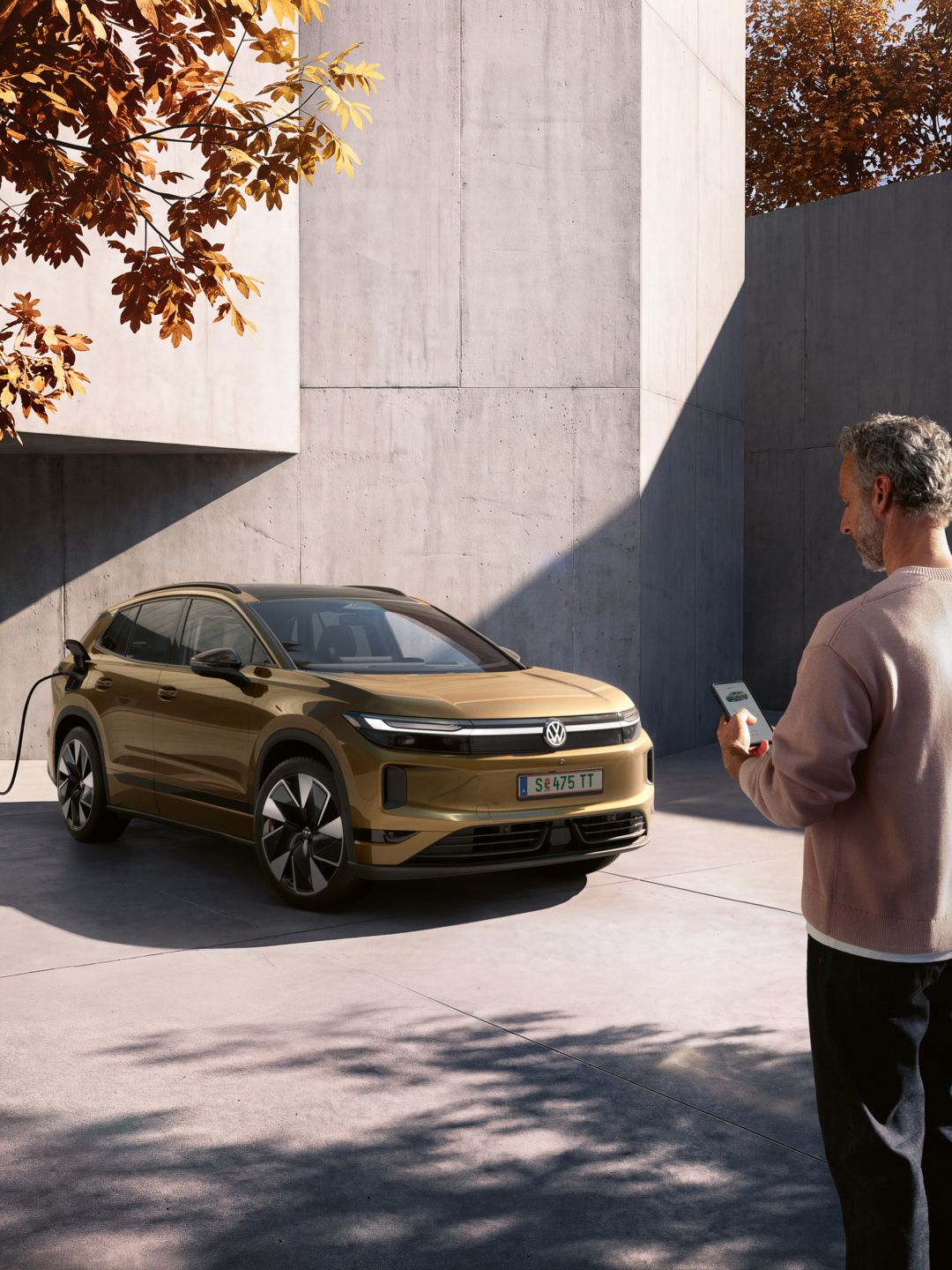 Ein Mann schaut auf das Handy in seiner Hand. Im Hintergrund lädt ein oliv-bronzefarbener VW ID.4 Reskin an einer Wallbox.