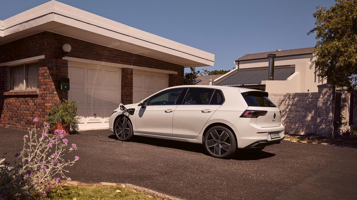 Ein weißer VW Golf eHybrid lädt an einer Ladestation vor einer Garage.