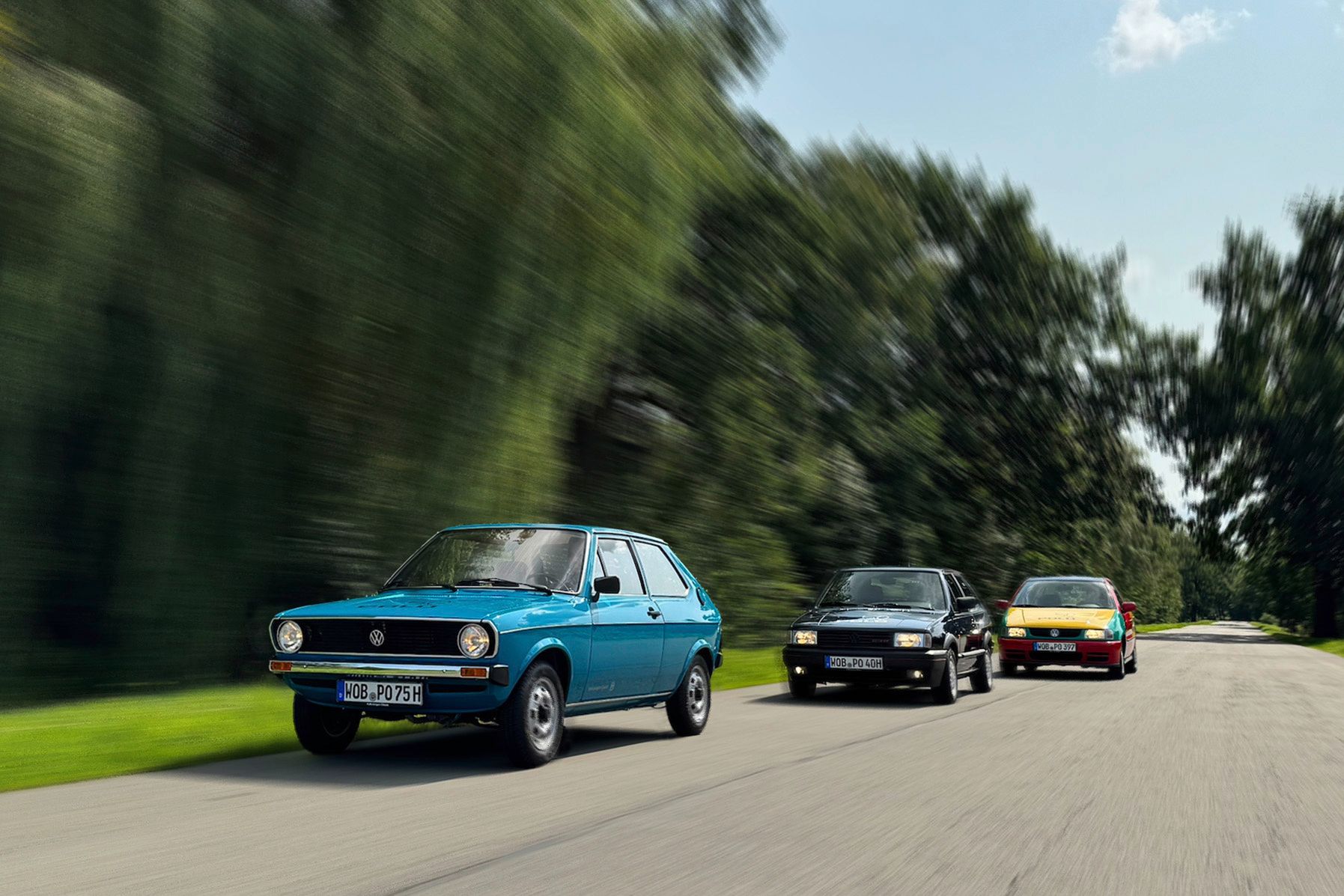 Drei VW Polos fahren hintereinander auf einer Landstraße durch eine grüne, waldige Landschaft. Das vordere Auto ist ein blauer Polo Mk1, das in der Mitte ein schwarzer Polo Mk2 und das hintere ein gelb-rot-grüner Polo Harlekin.