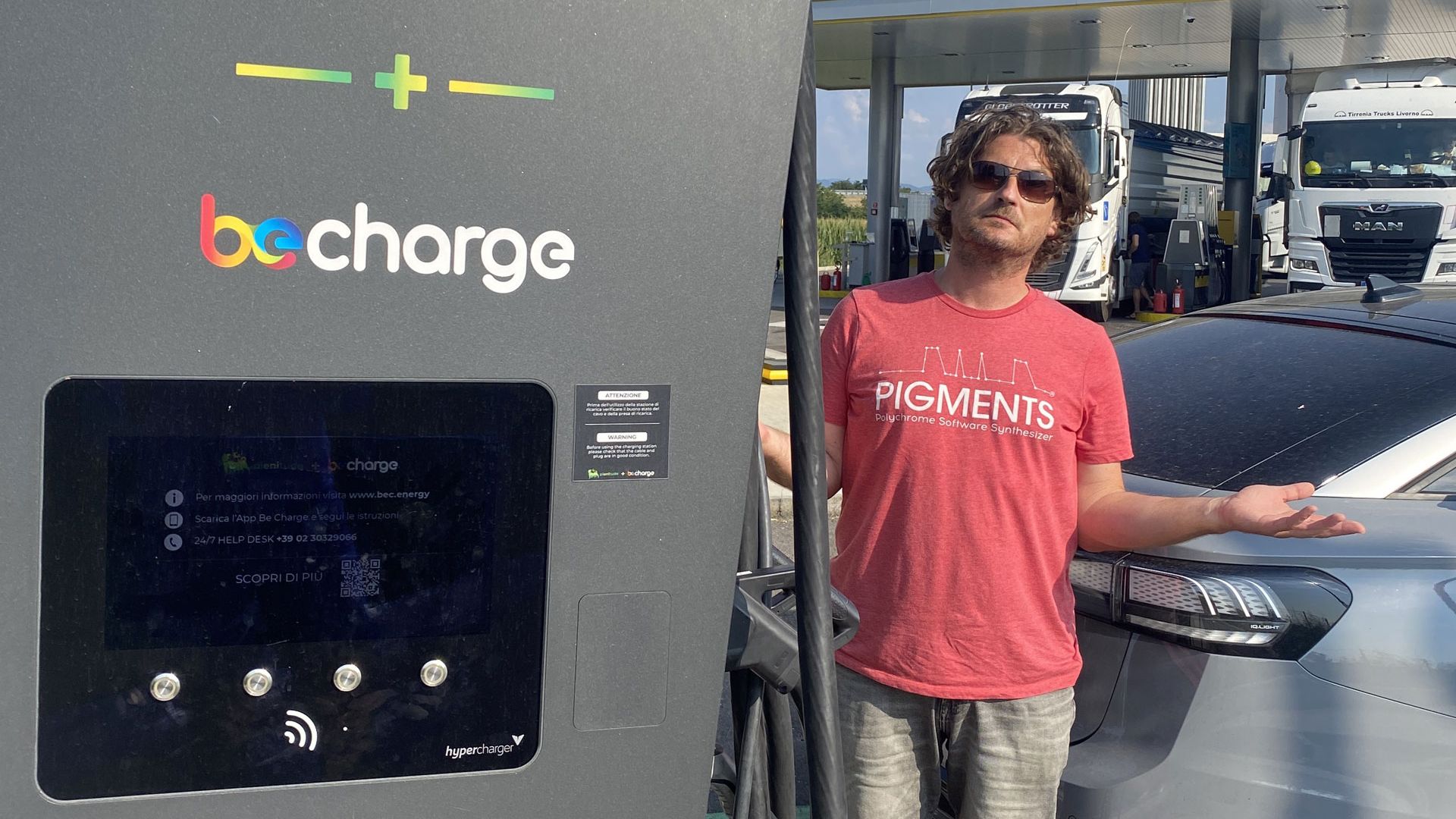 Ein Mann steht mit ausgestreckten Armen neben einer Elektroladestation und einem Elektroauto. Im Hintergrund sind mehrere Lastwagen zu sehen. Der Bildschirm der Ladestation zeigt Informationen zur Ladeleistung.