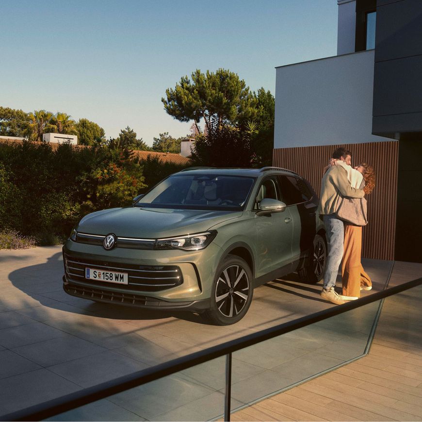 Eine Frau und ein Mann umarmen sich, neben ihnen steht ein VW Tiguan Elegance in der Farbe Cipressino-Grün Metallic