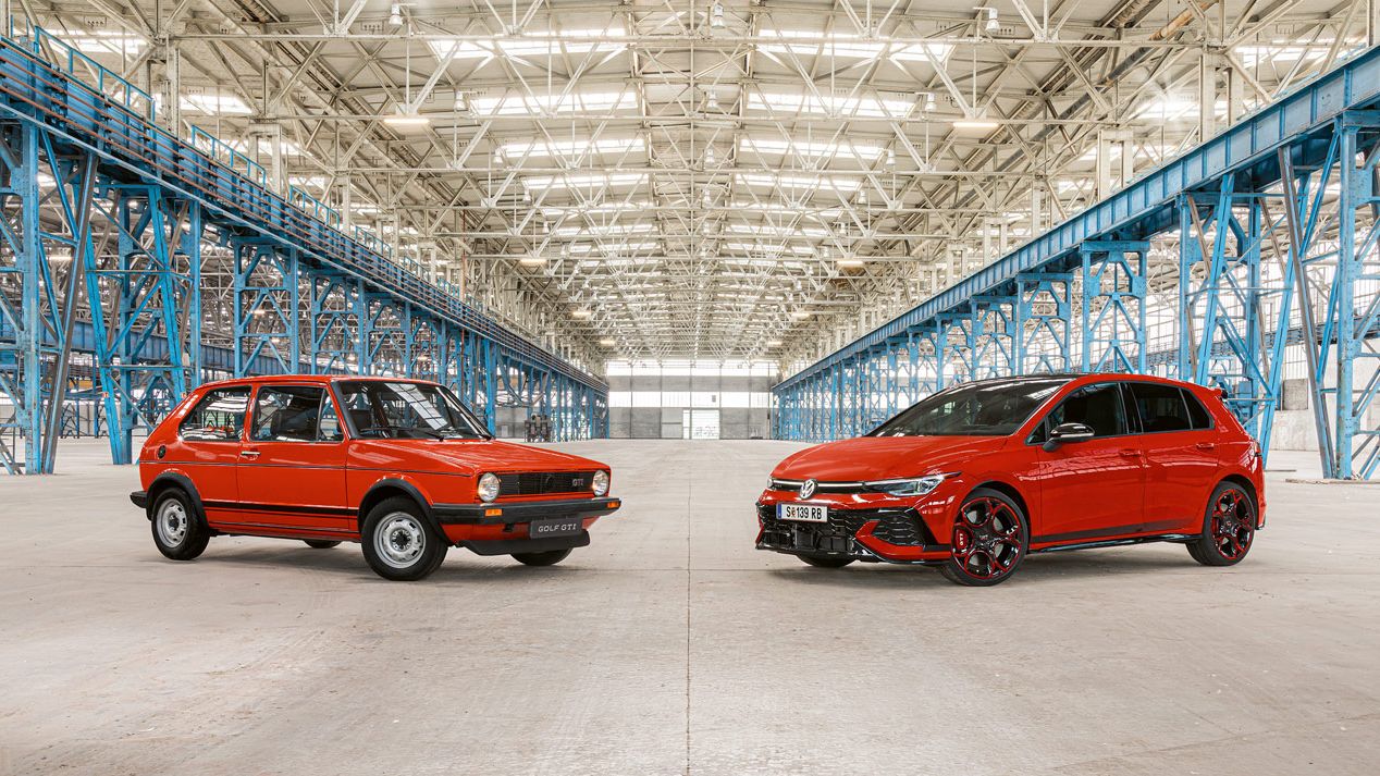 Zwei rote Golf GTI, das erste und das neuste Modell, stehen in einer großen Industriehalle mit vielen blauen Stahlträgern und einem hohen, hellen Dach.