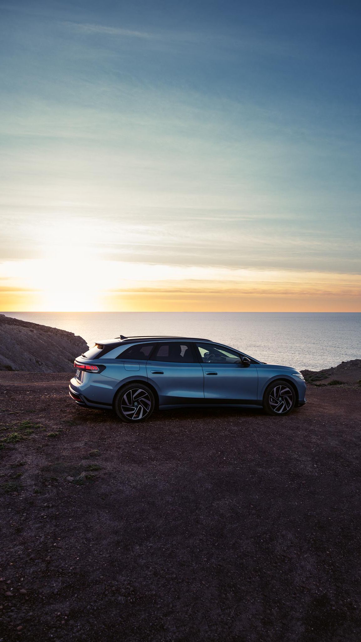 Ein blauer VW ID.7 Tourer steht oben auf einer Klippe im Sonnenuntergang, dahinter das Meer.
