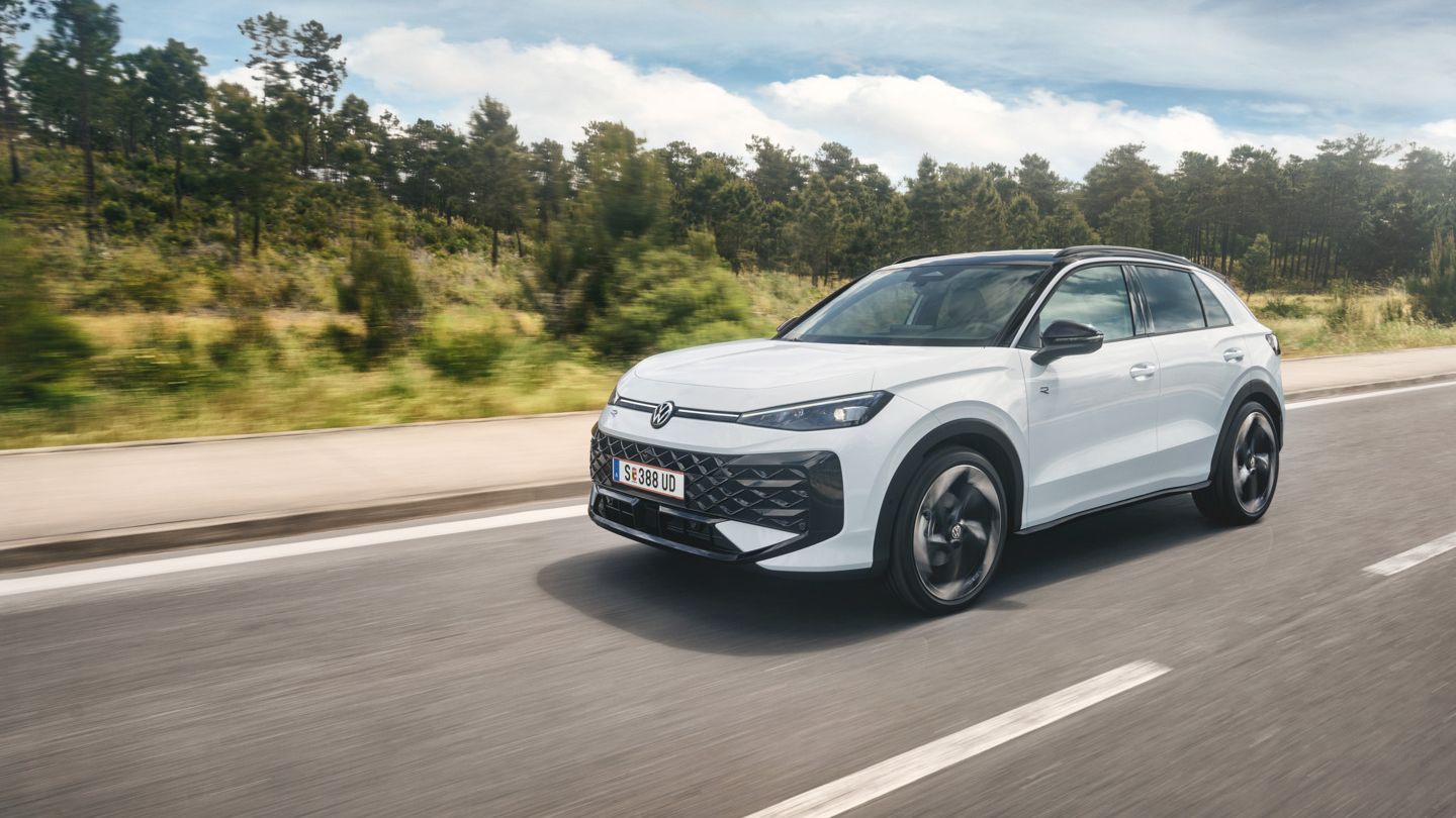 Ein weißer VW T-Roc Hybrid fährt auf einer Landstraße mit Wald im Hintergrund.