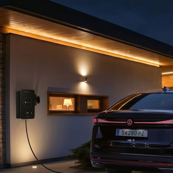 Ein schwarzes Elektroauto steht nachts in der Einfahrt eines modernen Hauses und ist an einer Wallbox zum Laden angeschlossen. Warmes, indirektes Licht an Fassade und Carport beleuchtet die Szene stimmungsvoll. Im Hintergrund sind Berge oder Hügel im Dämmerlicht zu erkennen.