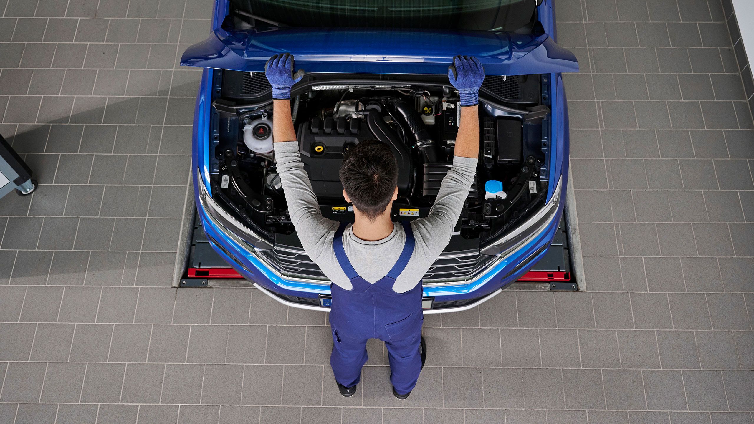 Mechaniker mit blauer Arbeitskleidung arbeitet unter der Motorhaube eines Autos. Der Motorraum ist deutlich sichtbar. Der Mechaniker steht auf grauem Fliesenboden.