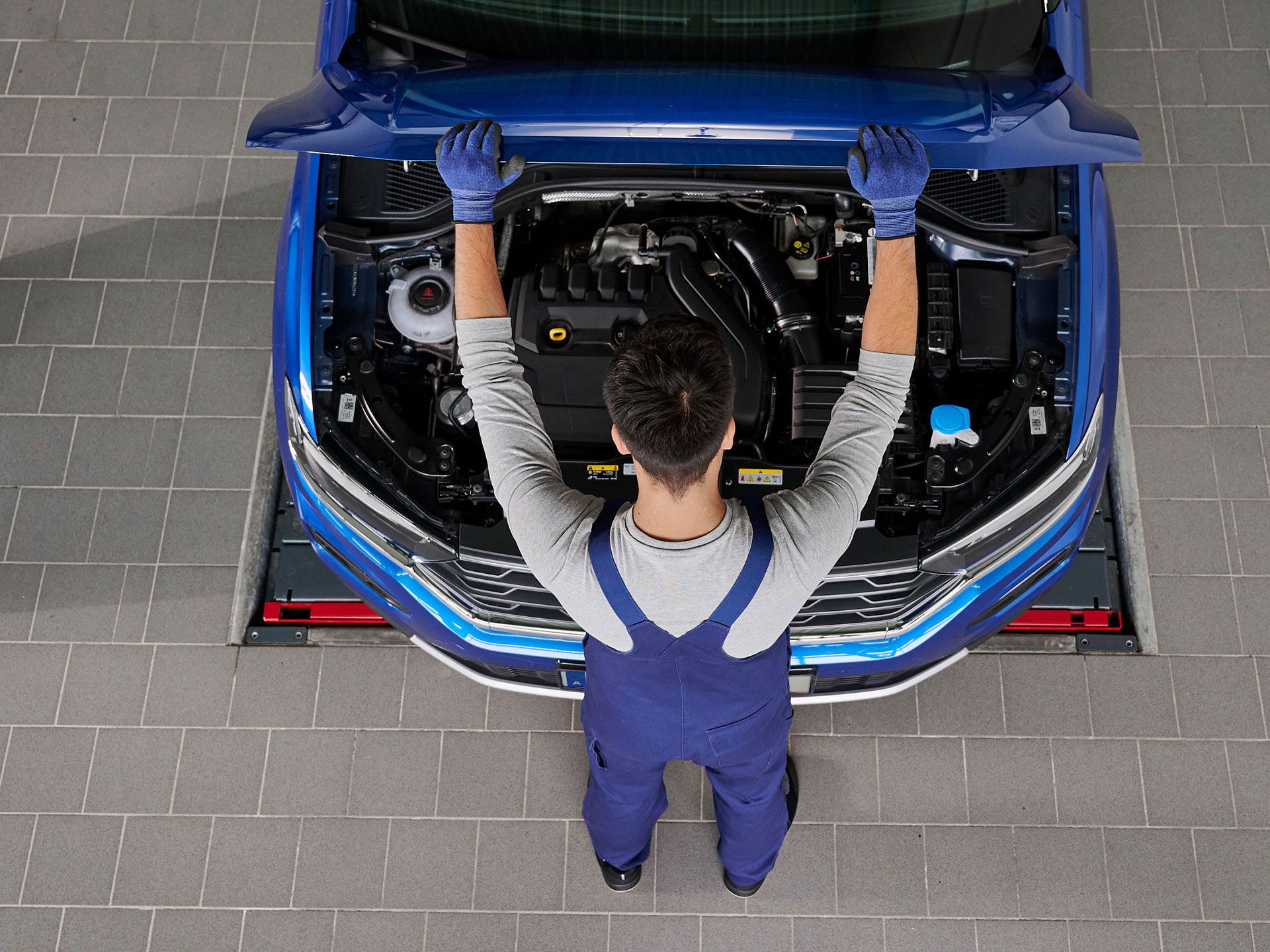 Mechaniker mit blauer Arbeitskleidung arbeitet unter der Motorhaube eines Autos. Der Motorraum ist deutlich sichtbar. Der Mechaniker steht auf grauem Fliesenboden.