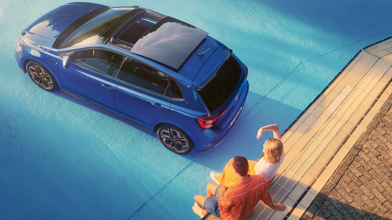  Blick von oben auf blauen VW Polo im Sonnenschein mit geöffnetem Panorama-Ausstell- /Schiebedach in einem leeren Pool. Zwei Personen sitzen am Rand des Pools.