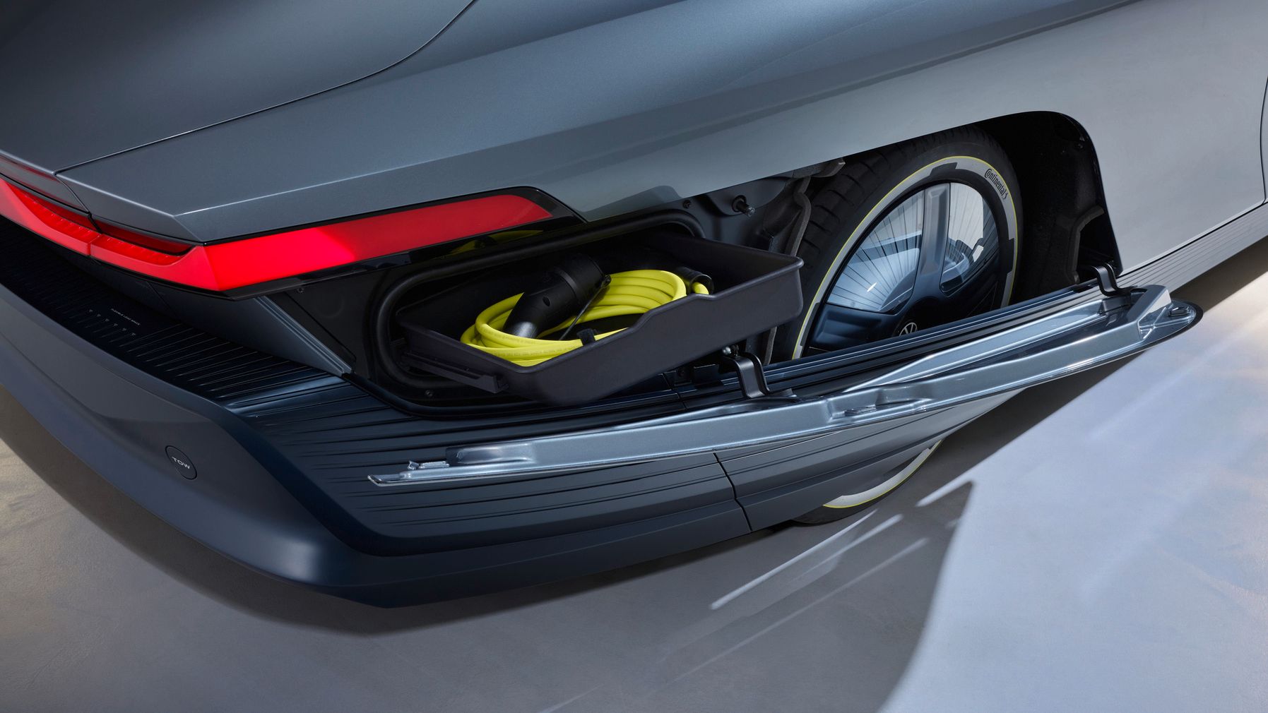 Rückansicht eines modernen Elektroautos, bei dem das seitliche Heckfach geöffnet ist. In dem Fach liegen ein gelb-schwarzes Ladekabel und Zubehör, dahinter ist ein Reifen sichtbar. Die Karosserie ist grau und wirkt futuristisch mit durchgehendem roten Rücklichtband.