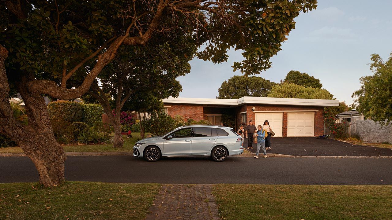 Weißer VW Golf Variant parkt in einer Wohnstraße mit Bäumen. Familie geht mit Gepäck darauf zu.