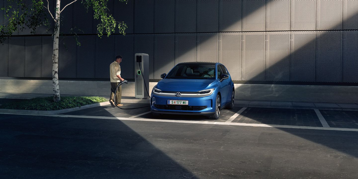 Ein blauer ID. Polo steht auf einem Parkplatz neben einer Ladesäule. Im Hintergrund sind ein Gebäude und ein Baum zu sehen.