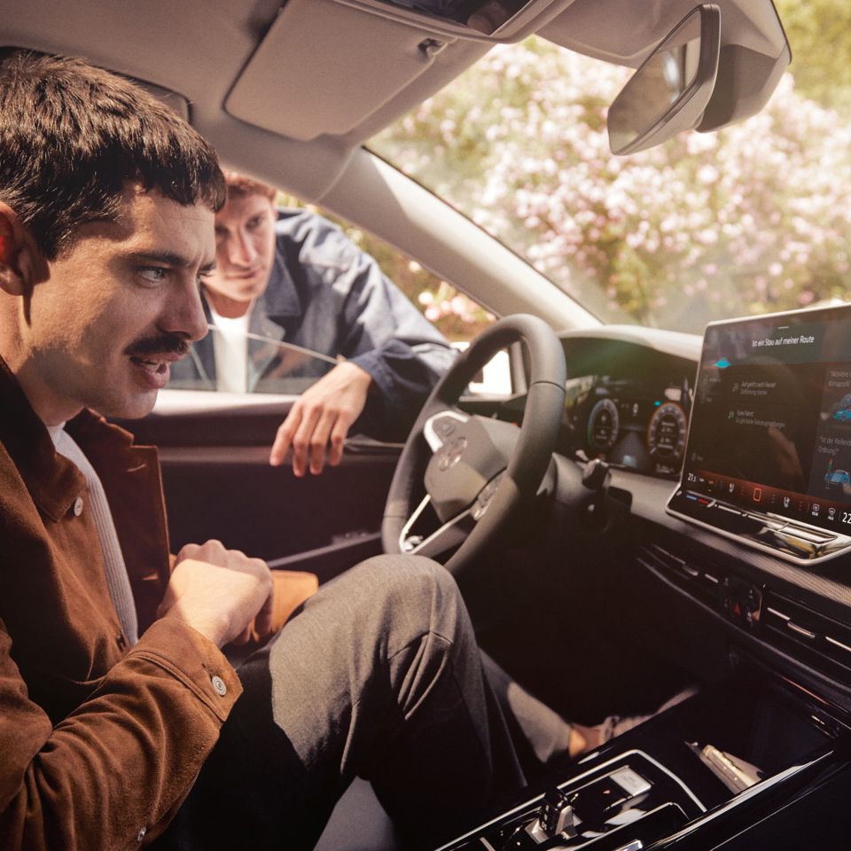 Ein Mann sitzt im neuen VW Golf und schaut auf das Infotainment-Display, ein anderer schaut durch das Fenster.