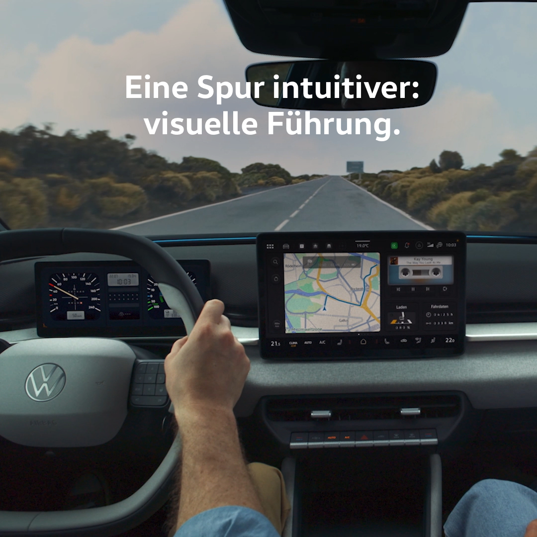 Blick aus dem Fahrzeuginnenraum auf eine gerade Landstraße, der Fahrer hält mit der rechten Hand das Lenkrad. Auf dem großen zentralen Display ist eine Navigationskarte zu sehen, links daneben das digitale Cockpit mit Tacho. Oben im Bild steht der Werbetext „Eine Spur intuitiver: visuelle Führung.“