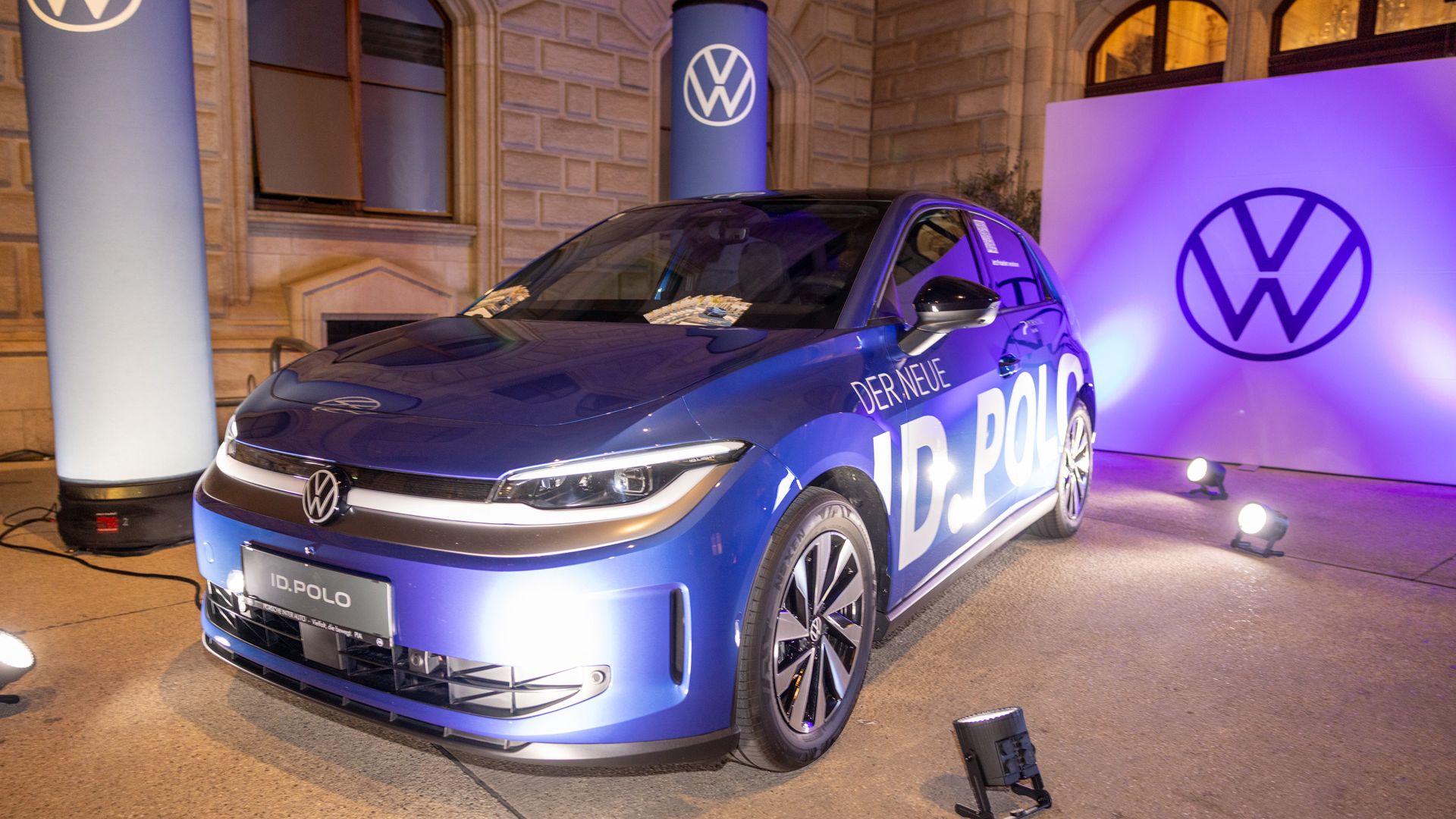 Blauer ID. Polo mit der Aufschrift „Der neue ID. Polo“ steht beleuchtet in einem Innenhof vor einem historischen Gebäude. Um das Auto herum sind Scheinwerfer, Werbestelen und eine Leinwand mit dem VW-Logo aufgebaut. Die Szene wirkt wie eine offizielle Fahrzeugpräsentation am Abend.