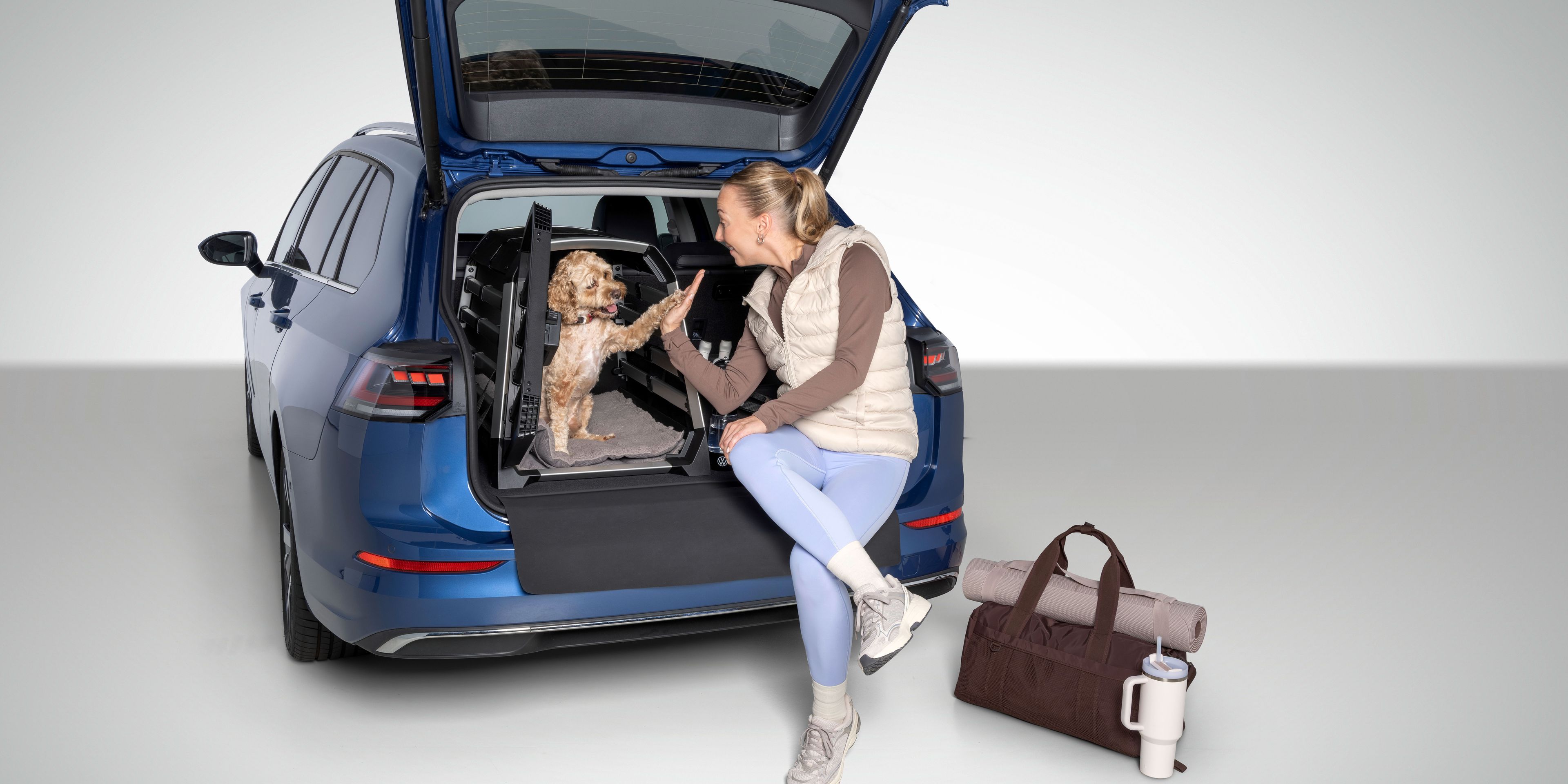Eine Frau sitzt auf der Ladekante eines blauen Volkswagen mit offener Heckklappe und gibt einem kleinen Hund im Kofferraum die Pfote.