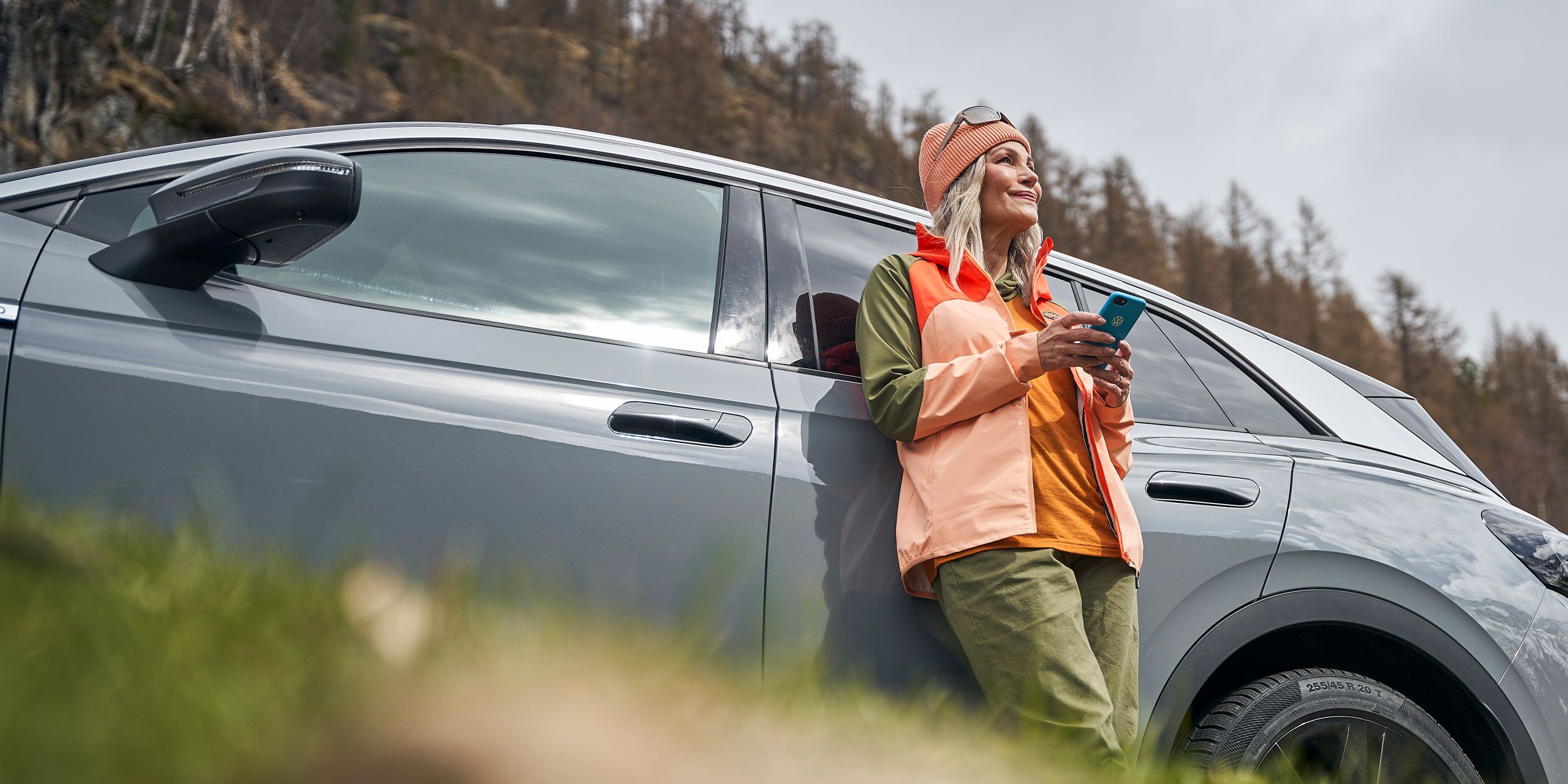 Eine Frau  mit Smartphone lehnt an einem Volkswagen und blickt in die Ferne